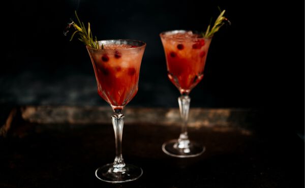 Two glasses of apple and cranberry Christmas spritz cocktails with rosemary sprigs on a dark wooden table.